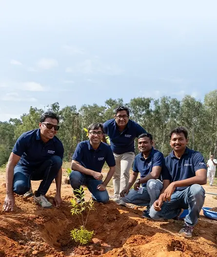 A group of five people in matching blue shirts pose near a newly planted sapling on a dirt patch. They smile and crouch down to show their participation in a tree-planting event, surrounded by trees in the background.